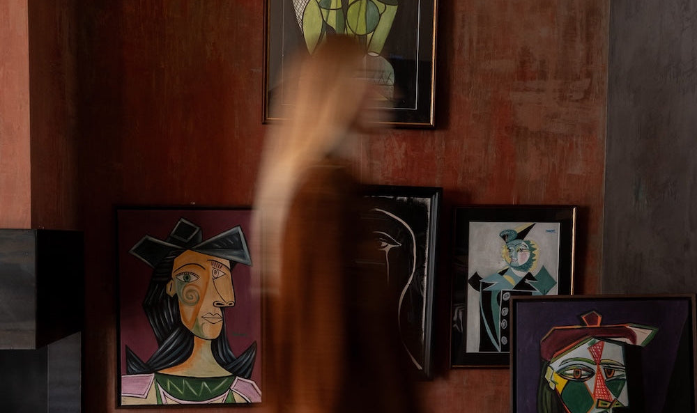 Eveline Kroese in a brown jacket and blue pants walks past a wall displaying multiple framed colorful Picassa paintings in a studio setting.