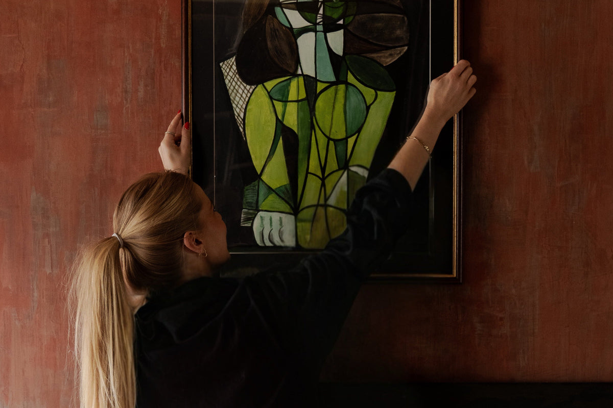 Eveline Kroese hangs a framed Picassa painting on a textured terracotta wall. The painting features a figure in vibrant green, black, and white with a dark brown frame.